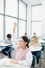 Fototapeta premium Vertical portrait of active smart twelve-year-old girl with dark hair sitting at school desk raising her hand in class, copy space