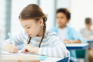 Horizontal medium close up portrait of Caucasian schoolgirl with two plaits sitting in modern classroom working on lesson task, copy space