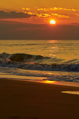 Colorful sunset at the tropical beach, sun behind clouds reflects on water and waves with foam hitting sand.