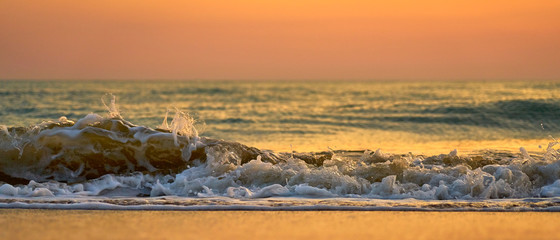 Colorful sunset at the tropical beach, sun behind clouds reflects on water and waves with foam hitting sand.