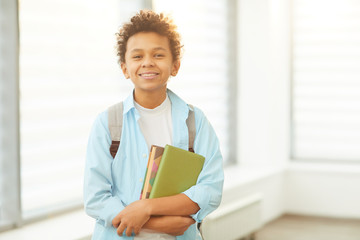 Horizontal medium portrait of young African American schoolboy wearing backpack holding books looking at camera smiling, copy space