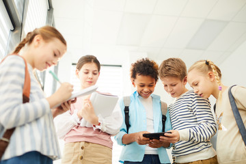 Cheerful boys and girls having fun standing together in school corridor watching something in Internet and chatting