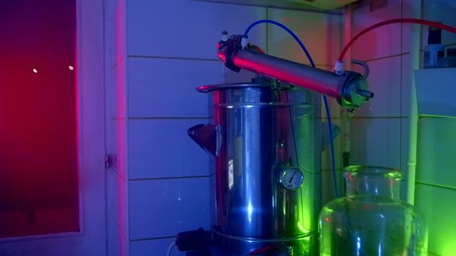 Making a strong drink at home. Moonshine still stands on a gas stove. The process of distilling alcohol in the kitchen. Moonshine slowly drips into a glass vessel. Green blue and red light. Close up