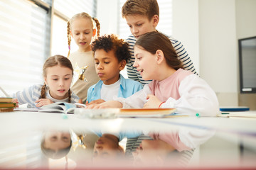 Group of young middle schoolers spending break time together in classroom looking at pictures and illustrations in book, horizontal shot - obrazy, fototapety, plakaty
