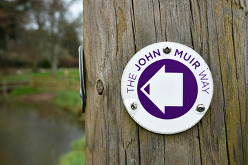 Sign for John Muir Way in Scotland with blurred background of grass and trees. Taken at East Linton. Sign is on wooden post and has a directional arrow.