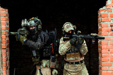 Two men in camouflage cloth and black uniform with machineguns stand beside brick wall. Soldiers...