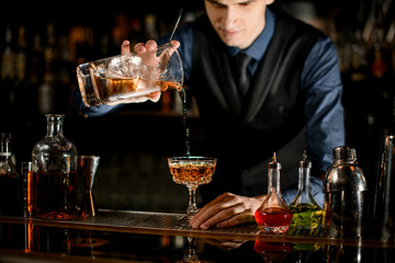 Young bartender pours a ready-made cold cocktail into wineglass.
