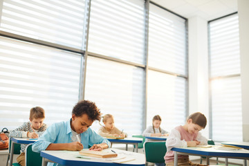 Horizontal shot of young middle school students sitting at desks in modern classroom doing lesson...