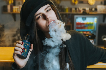 Young pretty woman in cap smoke an electronic cigarette at the vape shop. Hip-hop style. Closeup. - 328723969