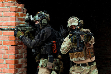 Two men in camouflage cloth and black uniform with machineguns stand beside brick wall. Soldiers with muchinegun aims aiming standing beside wall