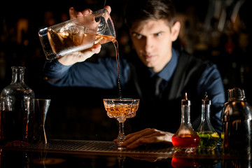 Bartender holds a glass with cocktail in his hand and pours it into wineglass.