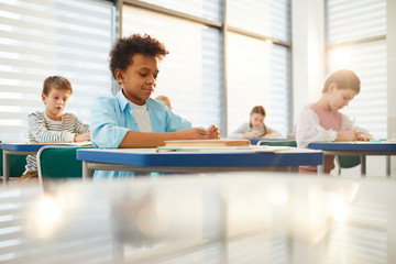 Horizontal shot of young middle schoolers wearing casual clothes sitting at desks in modern classroom doing lesson exercises, copy space - obrazy, fototapety, plakaty