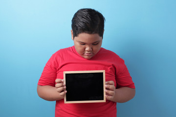 Sad fat Asian boy looking down while holding empty copy space blackboard