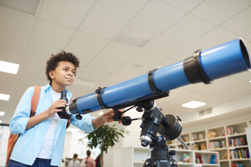 Horizontal medium low angle shot of African American middle schooler using modern telescope to at...