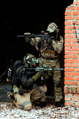 Two men in camouflage cloth green and black uniform with machineguns. Soldiers with muchinegun aims aiming in oposite sides, standing beside wall.