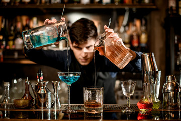 Young barman professionally pours two cocktails from one to another glass.