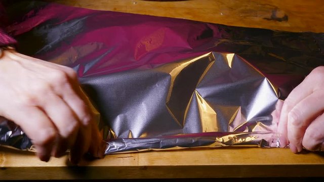 A woman&rsquo;s hands covering a piece of uncooked pink salmon, topped with sliced lemon, chopped garlic and red pepper, with silver foil, ready for baking.