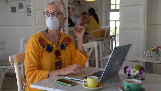 Tight shot of mature woman in cafe wearing protective face mask and looking away from her computer.