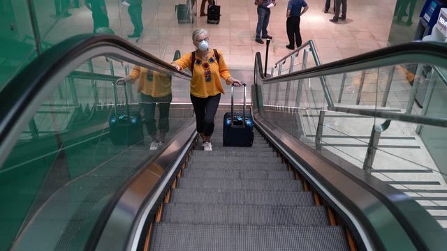 Distant mature woman wearing protective face mask riding escalator up with luggage in an airport.