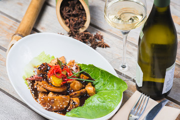 Sliced chicken fried in sweet and sour soy sauce with vegetables and herbs in a deep plate on a wooden table in a restaurant.