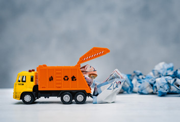 orange toy garbage truck unloads crumpled paper banknotes on a white table on a blue background.