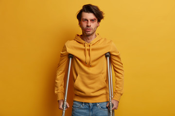 Health care concept. Disabled man with crutches being handicapped after tragic accident, has bruises and abrasion, disable to walk, isolated over yellow wall. Mobility assistance. Injured male