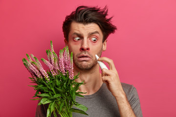 Shocked sick man holds nasal drops, has watery red eyes, suffers from allergy on pollen, has...