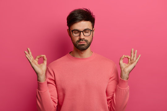 Patience, Calmness And Meditation Concept. Peaceful Relieved Bearded Young Man Practices Yoga Exercise, Keeps Hands In Zen Gesture, Closes Eyes, Poses Over Pink Background, Controls His Emotions