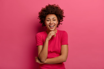 Fototapeta na wymiar Indoor shot of lovely woman giggles happily, looks straightly at camera, keeps hand under chin, listens hilarious joke, dressed in casual wear, poses against pink background. Emotions concept