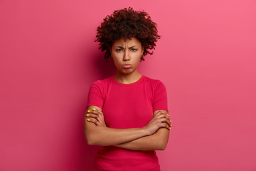 Resentful unhappy woman looks with sullen grimace, crosses arms over body, discontent with bad treatment, looks angrily at camera, wears red t shirt, poses over pink background. Negative emotions