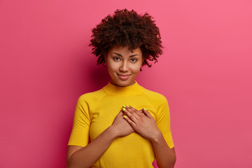Sign of affection and admiration. Pretty curly woman presses palms to heart, being grateful for gift, wears yellow t shirt, poses over rosy background, says you are always in my heart, smiles gently - obrazy, fototapety, plakaty