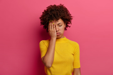 Fototapeta na wymiar Exhausted dark skinned young woman covers half of face, sighs from tiredness, has sleepy expression, closes eyes, wears yellow t shirt, poses over pink background. Female feels bored and tired