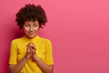 My plan is perfect. Pretty African American woman schemes something, steepls fingers and looks with cunning expression aside, smiles sly, poses over pink wall, copy space aside for your promo - obrazy, fototapety, plakaty