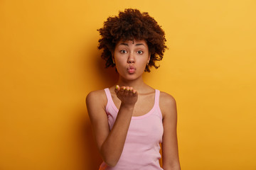 Romantic curly ethnic young woman keeps lips folded, sends air kiss, expresses love and flirts with boyfriend, gazes with tenderness, poses against yellow studio background. Body language concept