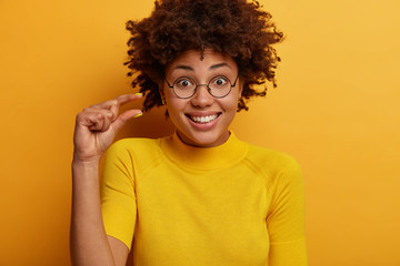 Little bit. Pretty smiling woman measures tiny invisble object, smiles gladfully, wears round glasses and casual t shirt, isolated on yellow background, tells about salary income or decreased price - obrazy, fototapety, plakaty