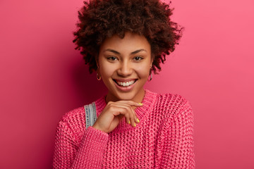Portrait of sensual curly woman smiles pleasantly, holds chin, expresses positive emotions, keeps smiling, shows white teeth, wears pink knitted sweater, has beauty face, healthy skin, curly hair