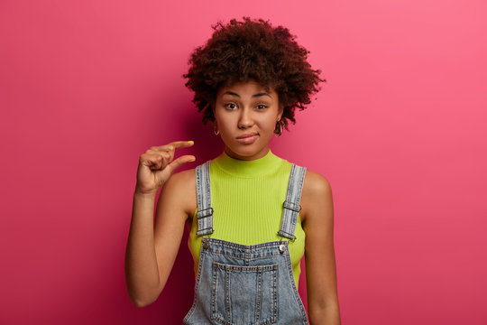 Displeased dark skinned woman has lack of time, shapes small or tiny object, wears casual clothes, explains how interested she was, has discontent expression, isolated over bright pink background