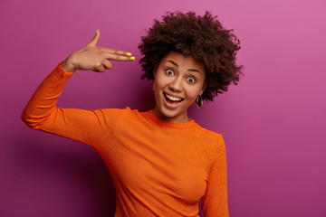 Horizontal shot of positive curly woman shoots in temples, makes suicide gesture, foolishes around, makes finger gun pistol, laughs happily, wears orange jumper, isolated on purple background
