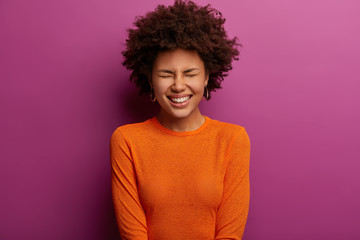 Ethnic overjoyed young woman laughs at something positive, closes eyes and giggles happily, dressed...