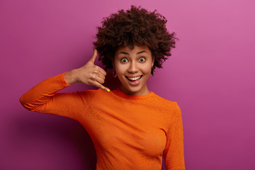 Fototapeta na wymiar Call me later. Pleased curly young woman makes phone gesture, pretends mobile communication, smiles happily, wears casual orange jumper, isolated over purple studio wall. Body language concept