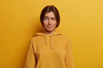 Beautiful dark haired young European woman presses lips, raises eyebrows, has worried face expression, wears casual hoodie, poses against yellow background, has embarrassed look. Monochrome shot.