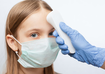 Doctor using infrared thermometer checking body temperature of girl wearing surgical mask -...