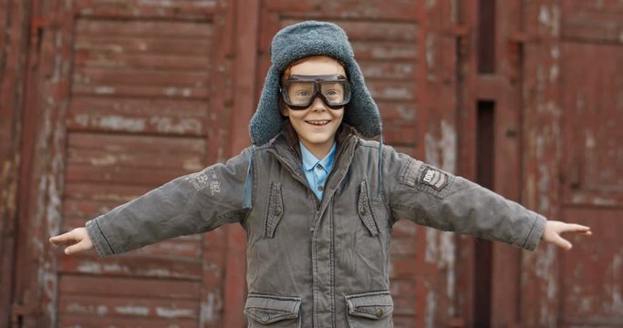 Joyful Caucasian cute little boy with red hair, glasses and hat smiling and staying outdoor while playing in aviator. Small kid dreaming to be pilot when growing up.