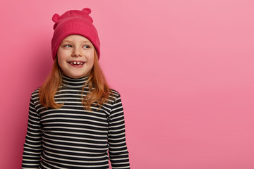 Redhead positive girl wears hat and striped poloneck, looks aside, notices something amused, remembers spending nice time with parents, poses over pastel rosy background, has lovely expression