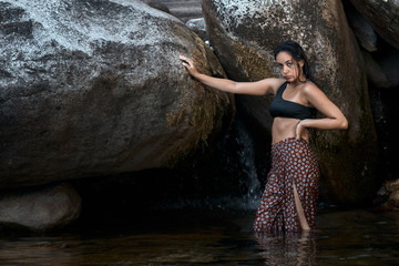 Joven morena de pelo de pie posando junto a las rocas en las montañas mojada dentro del agua vestida.