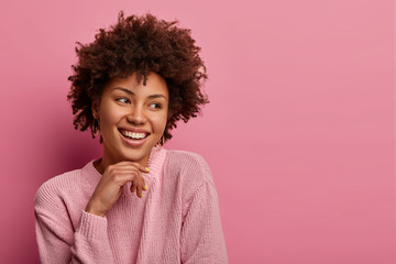 Headshot of pretty charming curly Afro woman poses joyful and looks aside, keeps hand under chin,...