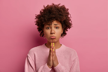 Hopeful sad uneasy curly woman beggs for forgiveness, purses lips and keeps palms in praying gesture, asks mercy and says please, needs your help, wears casual jumper, poses over pink background