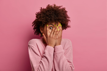 Frightened woman covers face with palms, peeks through fingers, sees something horrible and unexpected, hides from all difficulties, intrigued what happening, wears jumper in one tone with background