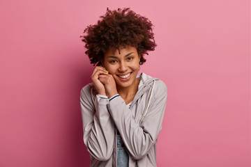Image of positive charming millennial girl keeps hands together near face, looks with tender expression at camera, hears something pleasant, poses over pink background. Positive feelings concept