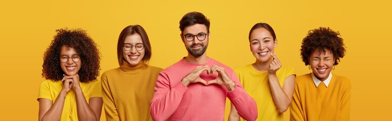 Pleased bearded adult man makes heart gesture over chest, poses between four women with cheerful...